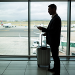 Man with a luggage standing on an airport lounge with the runway at the background.